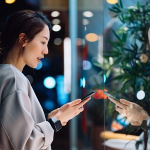 Smiling young Asian woman using smartphone while shopping in the city, standing outside a boutique shop window in the evening