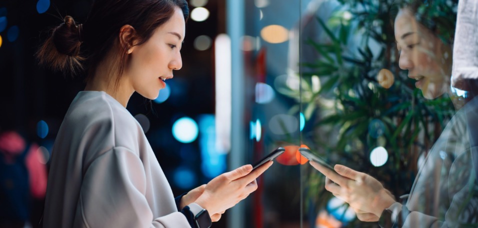 Smiling young Asian woman using smartphone while shopping in the city, standing outside a boutique shop window in the evening